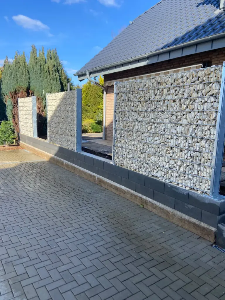 Sichtschutz aus Gabionen auf einem Sockel aus Betonsteinen, davor Pflaster im Fischgrätverband