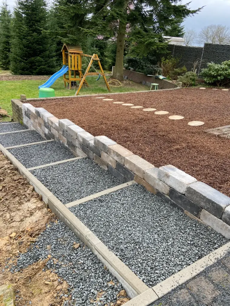 Gartentreppe mit Schotterstufen und Einfassung, daneben eine Mauer aus Blocksteinen und eine Fläche mit Rindenmulch